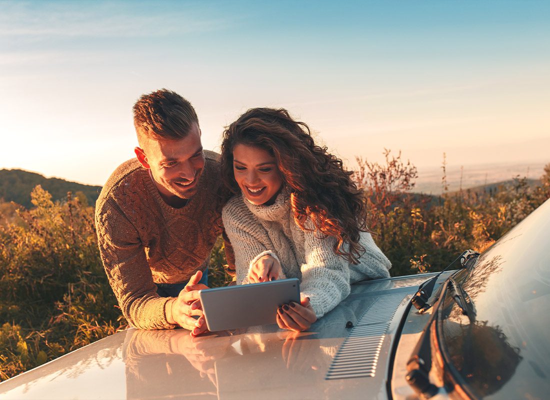 Service Center - A Young Man and a Young Woman Smiling Down at a Tablet While Resting on Their Car